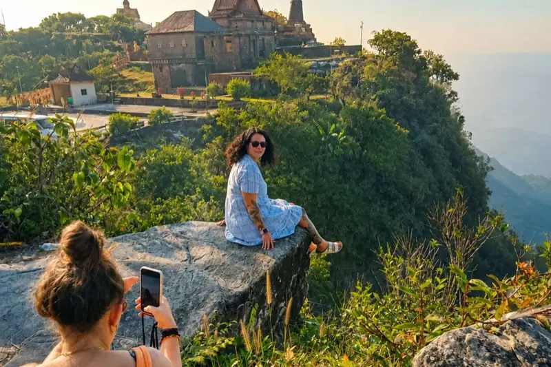 Eine Reisende sitzt auf einem Felsvorsprung hoch auf dem Bokor-Berg, eine Freundin fotografiert sie, dahinter eine Pagode